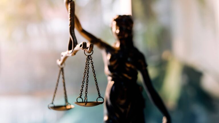 Close-up of a bronze Lady Justice statue holding balanced scales, with a blurred background and sunlight shining from the left.