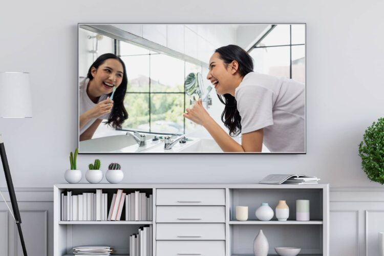 A TV on a living room wall displays a woman smiling and brushing her teeth in front of a bathroom mirror. Shelves, books, and plants are arranged below the TV.
