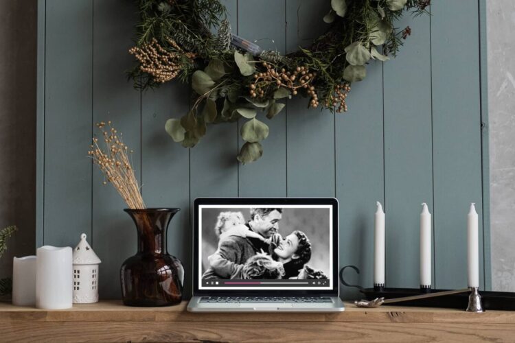 A laptop on a wooden table displays a black-and-white movie scene. The table also holds candles, vases, and decor, with a wreath hanging on the blue wall behind.