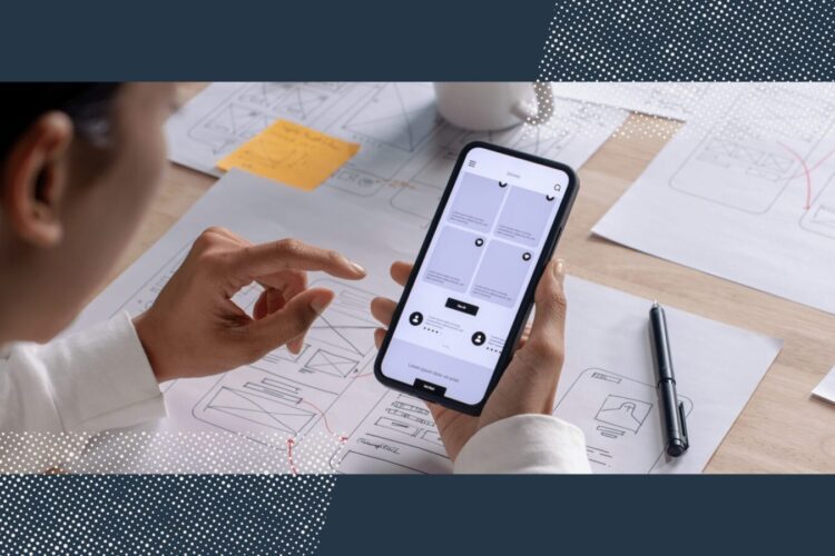A person uses a smartphone to view a wireframe app design, with printed wireframes, a pen, and notes on a desk in the background.