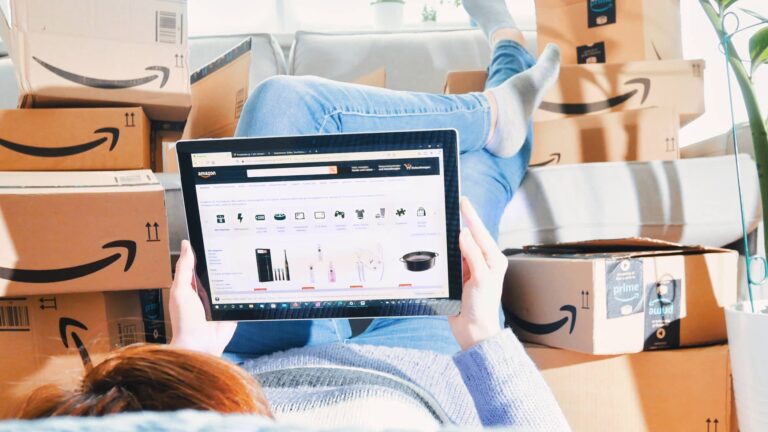 A person lies on a couch surrounded by Amazon boxes, browsing online shopping on a tablet.