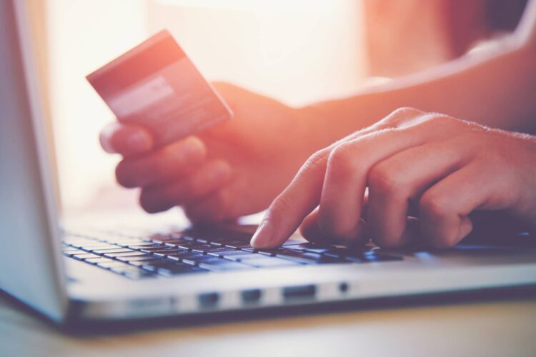 Person holding a credit card in one hand and typing on a laptop keyboard with the other, suggesting online shopping or payment.