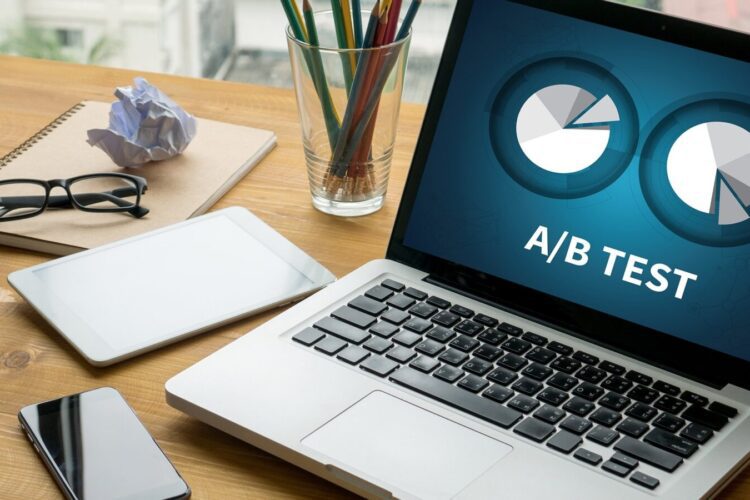 A laptop displaying an A/B test graphic sits on a wooden desk with a tablet, smartphone, glasses, crumpled paper, and a notebook with colored pencils.