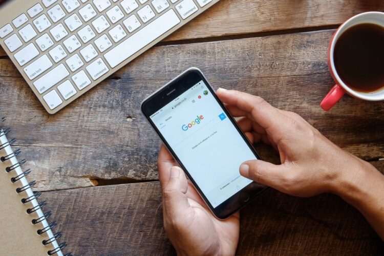 Person holding a smartphone with Google search open, next to a keyboard, notebook, compass, and a cup of coffee on a wooden desk.