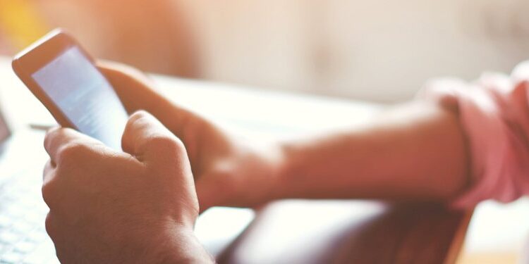 Person using a smartphone while sitting at a desk with an open laptop, with sunlight streaming in from the left side of the image.