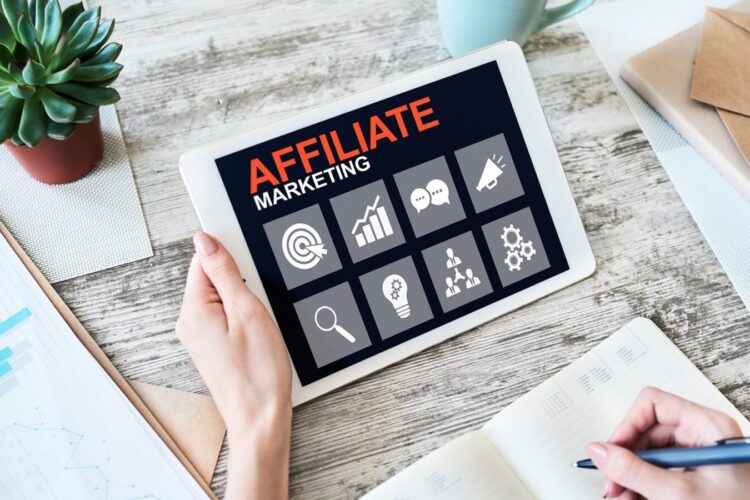Person holding a tablet displaying "Affiliate Marketing" with related icons, next to a notebook, pen, and a potted plant on a wooden desk.