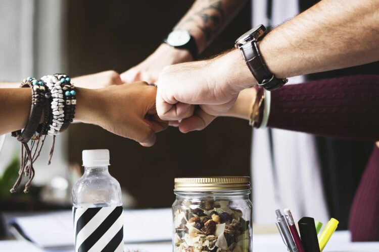 Four people join fists over a desk with a jar of mixed nuts, a water bottle, and office supplies in view.