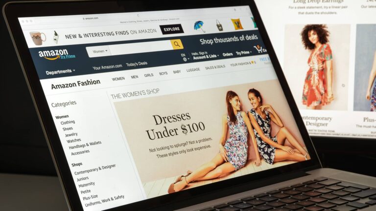 Laptop screen displays Amazon Fashion homepage, highlighting "Dresses Under $100" with images of two women in dresses and a navigation menu on the left.