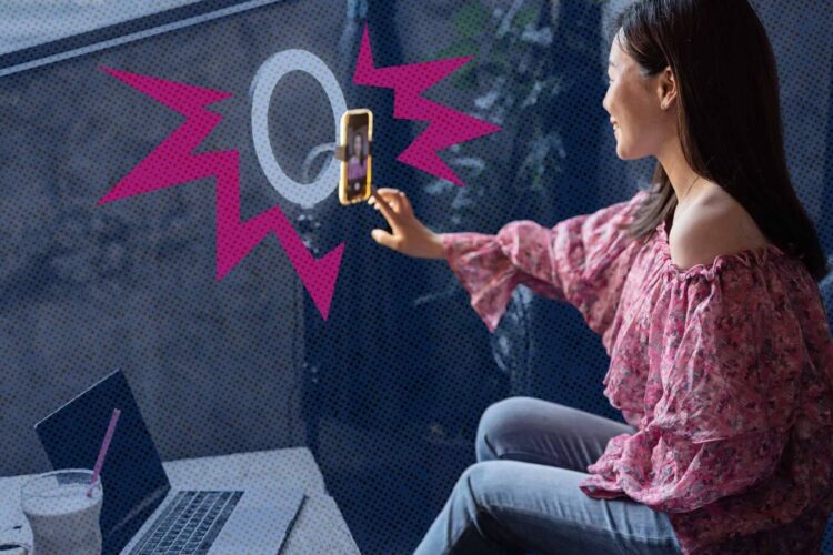 A woman in a pink top sits by a window, holding a smartphone on a ring light stand, with a laptop open on the table in front of her.