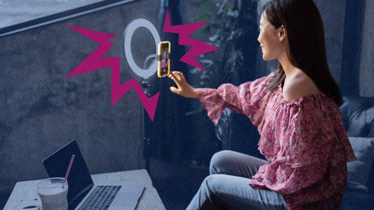A woman in a pink top sits by a window, holding a smartphone on a ring light stand, with a laptop open on the table in front of her.