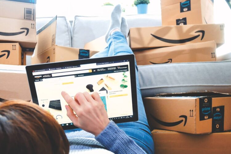 Person lying on a couch using a tablet to shop on Amazon, surrounded by multiple Amazon delivery boxes.