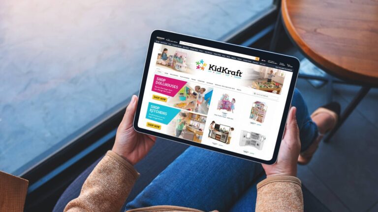 Person holding a tablet displaying the KidKraft website, which features sections for dollhouses, play kitchens, and other children's toys.