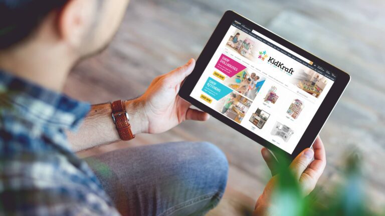 A person holds a tablet displaying the KidKraft website, which features images and links for toys, sales, and playhouse products.