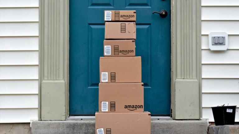 Six Amazon shipping boxes stacked in a vertical column on the doorstep of a house with a blue door.