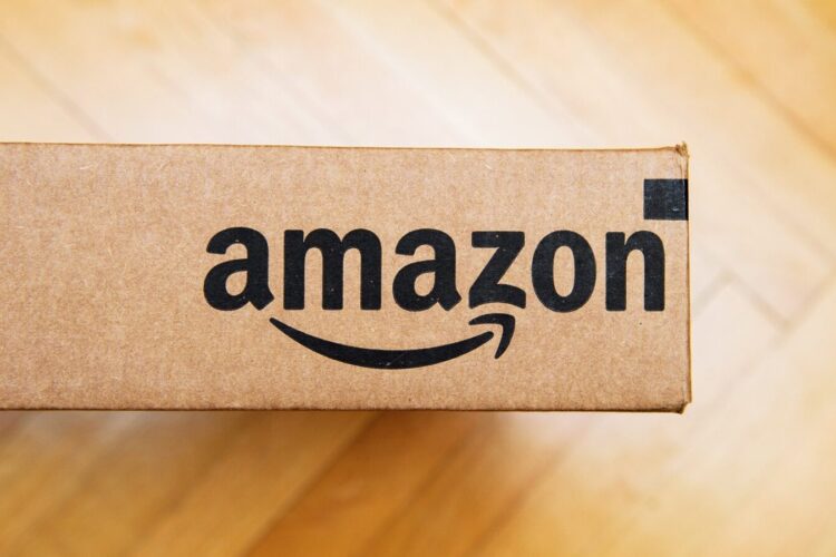 A brown cardboard box with the Amazon logo printed in black, placed on a light wood floor.