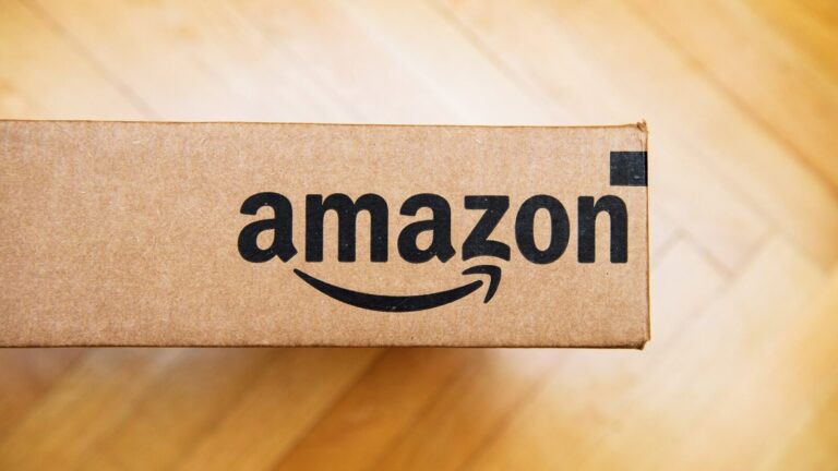 A brown cardboard box with the Amazon logo printed in black, placed on a light wood floor.