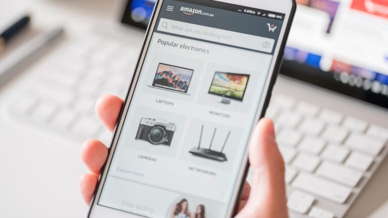 A person holds a smartphone displaying the Amazon website, showing popular electronics categories such as laptops, monitors, cameras, and networking. A laptop is visible in the background.