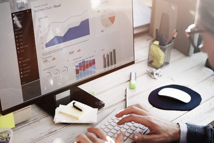 Person at desk working on a computer displaying business charts and graphs, surrounded by office supplies, a cup, and a keyboard.