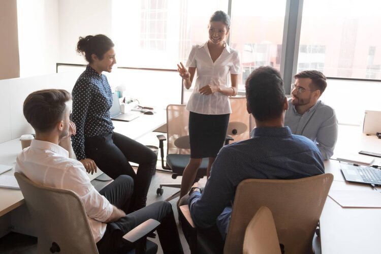 Five people in business attire have a discussion in a modern office, seated in a semi-circle around a woman who is standing and gesturing while speaking.