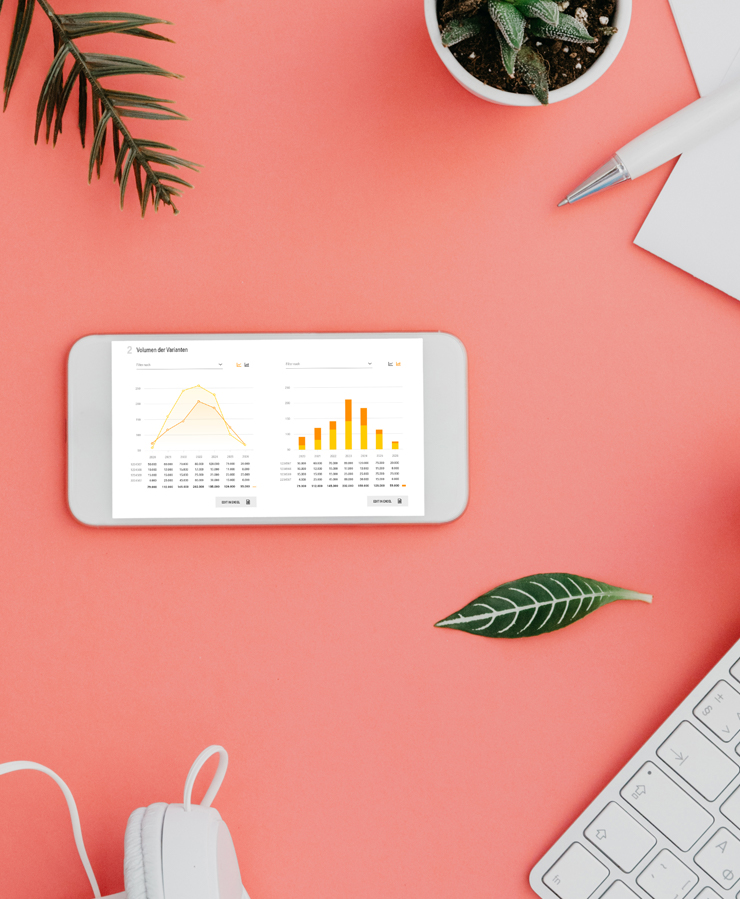 A smartphone displaying graphs lies on a coral desk beside a keyboard, pen, notebook, headphones, leaves, and a potted succulent.