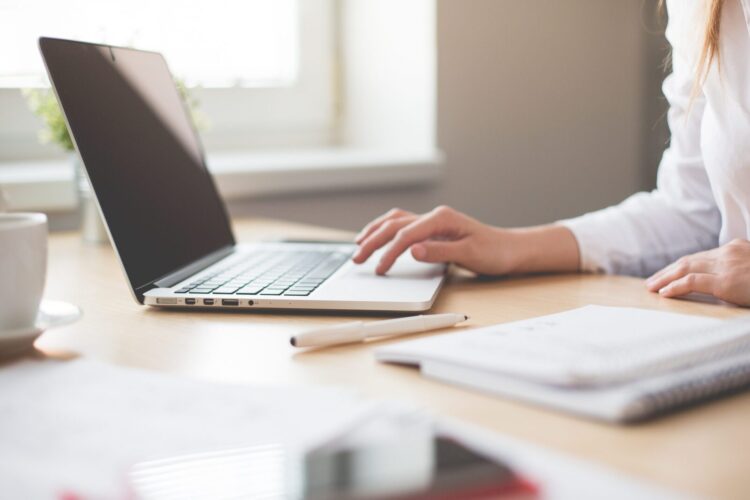 Person working at a desk using a laptop; a notebook, pen, and coffee cup are also on the desk, with natural light coming through a nearby window.