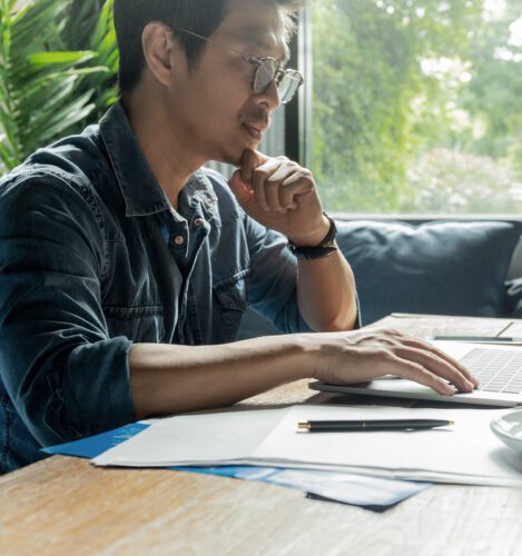 A person sits at a desk using a laptop, with papers and a pen nearby, in a room with large windows and natural light.