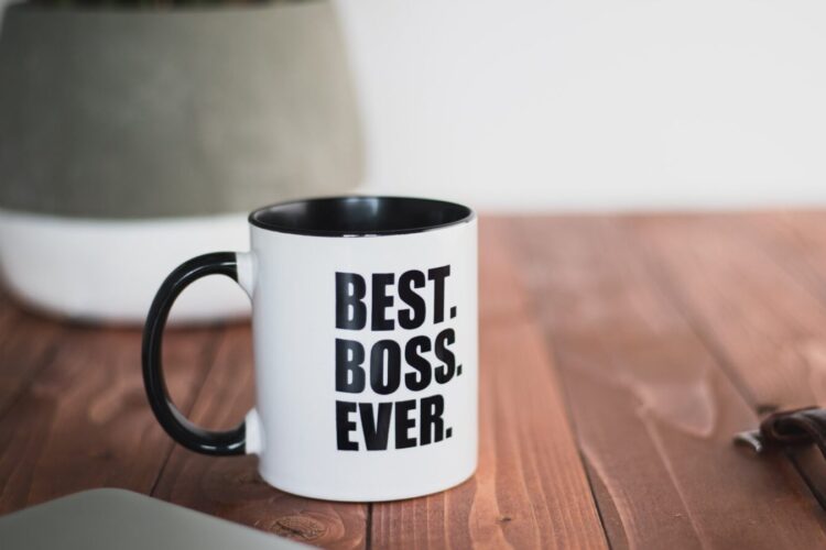 A white coffee mug with black handle and rim reads "BEST. BOSS. EVER." in bold letters, sitting on a wooden table next to a plant pot.