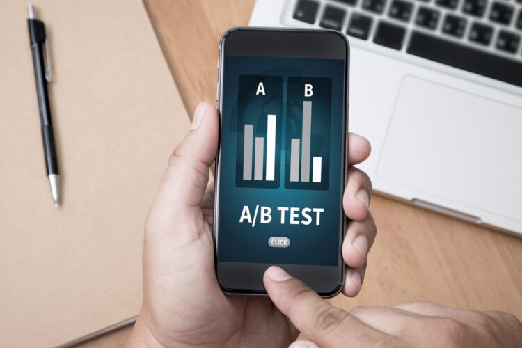 Person holding a smartphone displaying an A/B test comparison chart, with a laptop, pen, and notebook on a desk in the background.