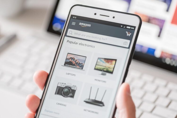A person holds a smartphone displaying the Amazon app's Popular Electronics page, with categories for laptops, monitors, cameras, and routers visible. A blurred laptop screen is in the background.