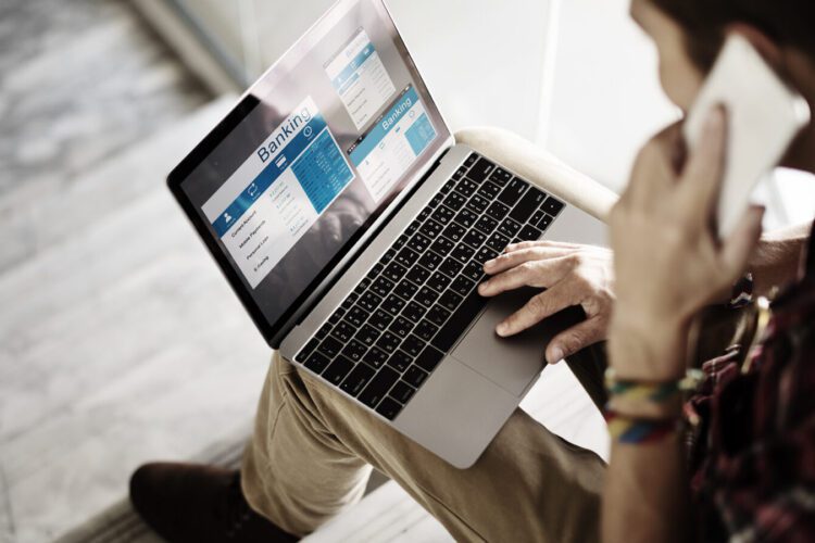 Person sitting and using a laptop displaying online banking screens while holding a smartphone to their ear.
