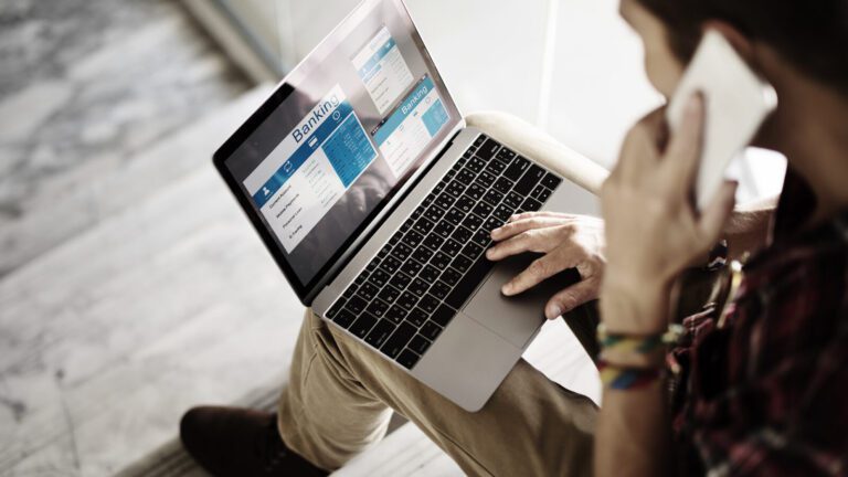 Person sitting and using a laptop displaying online banking screens while holding a smartphone to their ear.