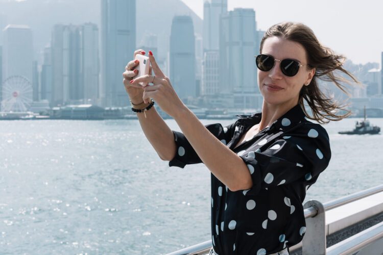 A woman wearing sunglasses takes a selfie by the waterfront with a city skyline and ferris wheel in the background.