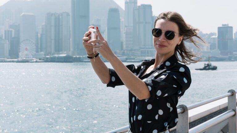 A woman wearing sunglasses takes a selfie by the waterfront with a city skyline and ferris wheel in the background.