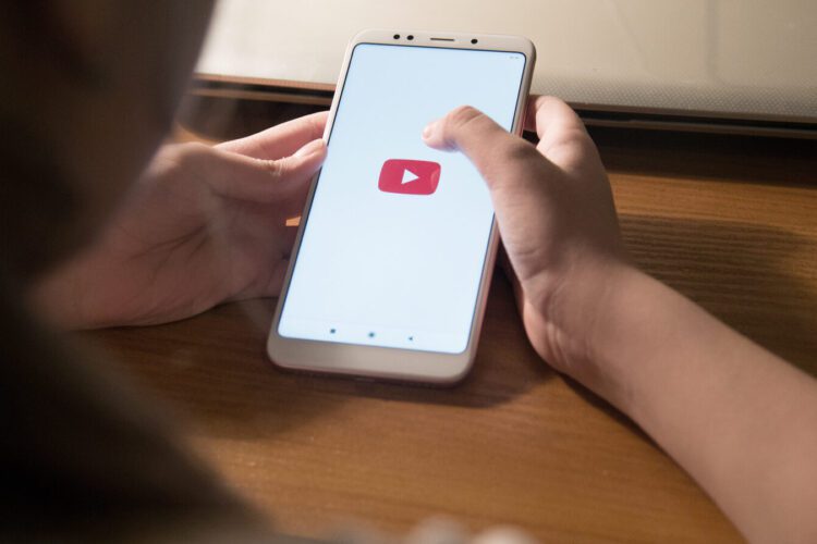 Person holding a smartphone displaying the YouTube app logo on the screen, seated at a wooden desk.