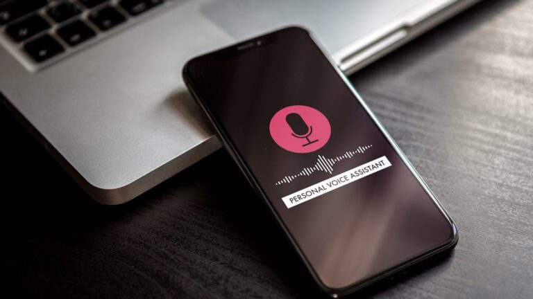 A smartphone with a personal voice assistant interface is on a dark desk next to a laptop keyboard.