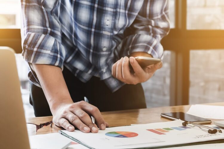 Person in plaid shirt stands at desk holding a smartphone, reviewing charts and graphs on paper next to a laptop and other office items.