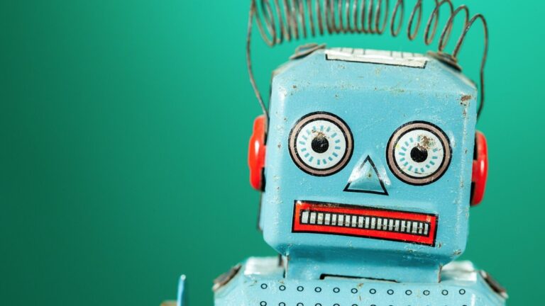 Close-up of a vintage blue toy robot with red ears, a coiled spring antenna, and a painted face, set against a plain green background.