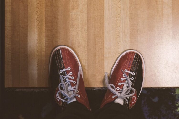 A person wearing red and black bowling shoes stands on a wooden floor, viewed from above.