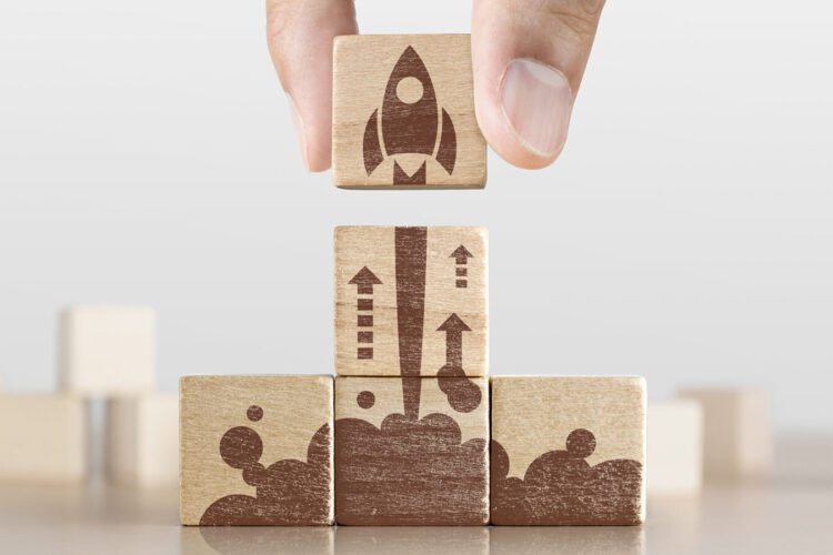 A hand places a wooden block with a rocket icon on top of three stacked blocks showing a launch sequence with arrows and smoke.