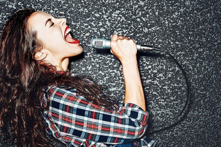 Woman with long curly hair and red lipstick singing passionately into a microphone against a textured dark background, wearing a plaid shirt.