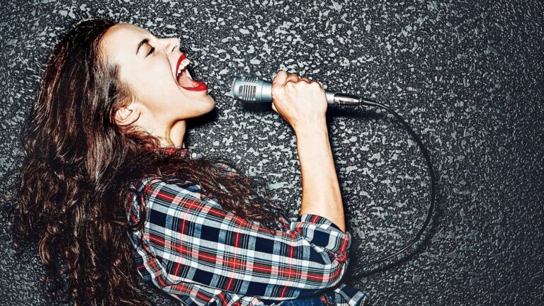 Woman with long curly hair and red lipstick singing passionately into a microphone against a textured dark background, wearing a plaid shirt.