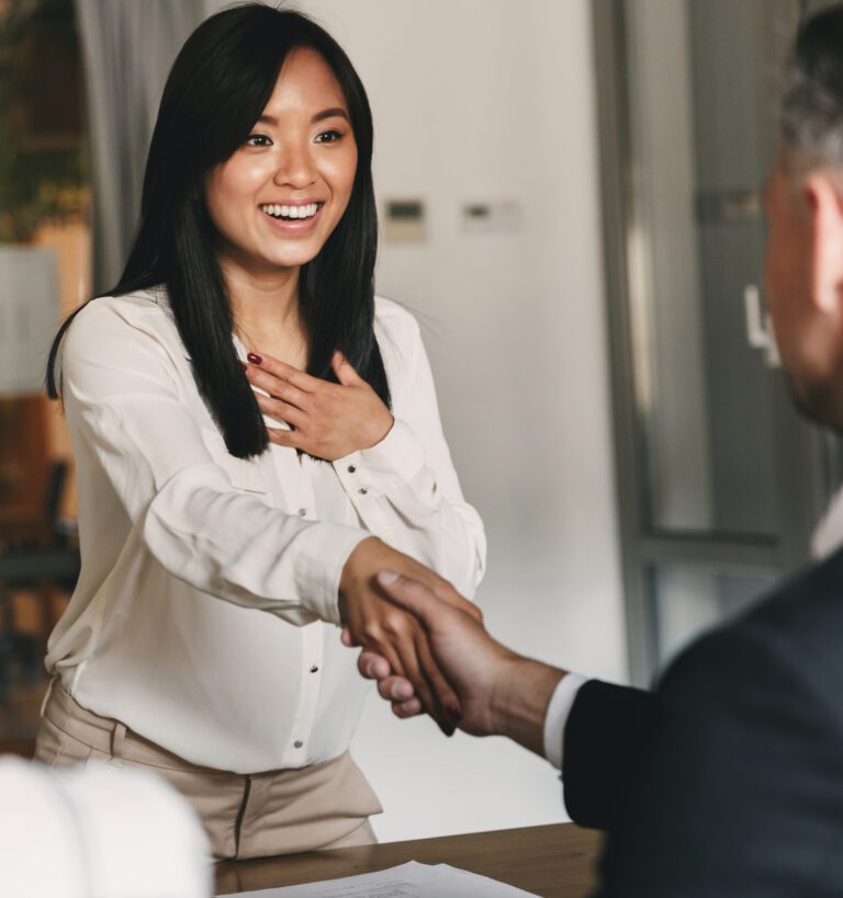 A woman smiles and shakes hands with a man across a desk in an office setting, appearing happy and engaged.
