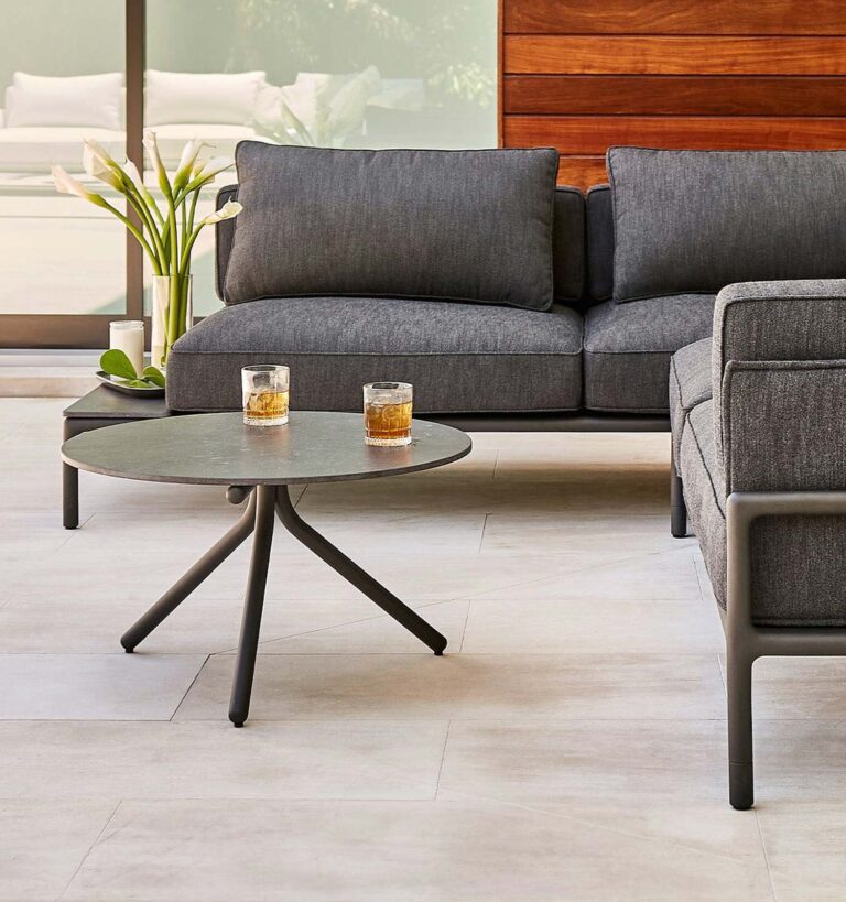 A modern outdoor patio with a gray sofa, a matching armchair, a round black coffee table, and two glasses with drinks on the table. White lilies are in a vase in the background.