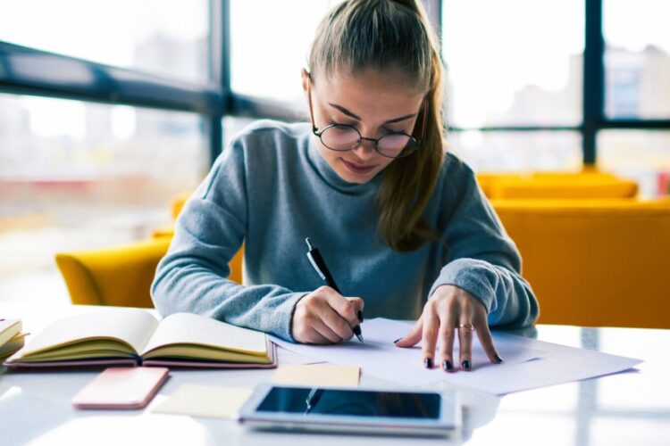 A young woman wearing glasses writes on paper at a table with notebooks, a phone, and a tablet in front of her, with large windows and yellow chairs in the background.