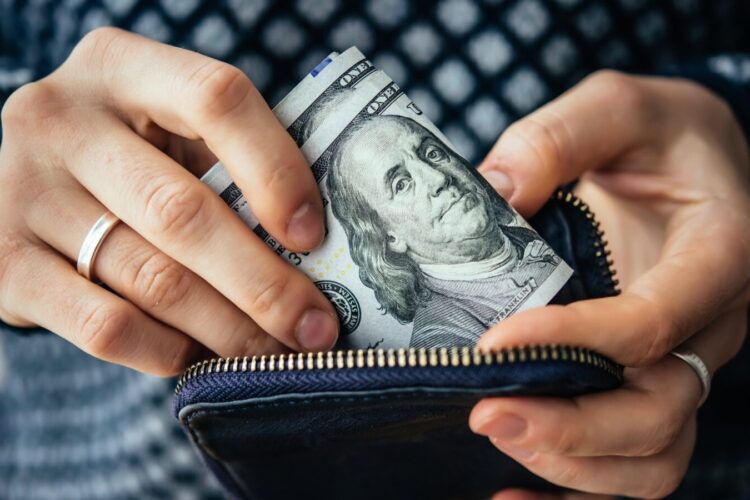 A person holding a wallet and pulling out a roll of U.S. hundred dollar bills.