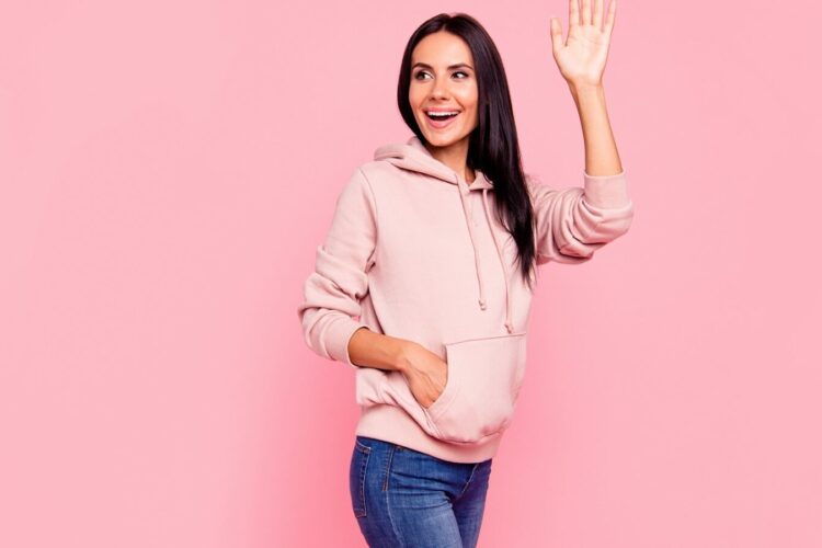 Woman with long dark hair wearing a light pink hoodie and blue jeans stands against a pink background, smiling and raising her right hand in a wave.