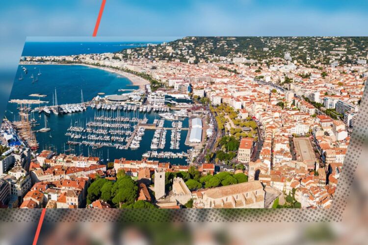 Aerial view of a coastal city with a marina filled with boats, red-roofed buildings, and hills in the background.