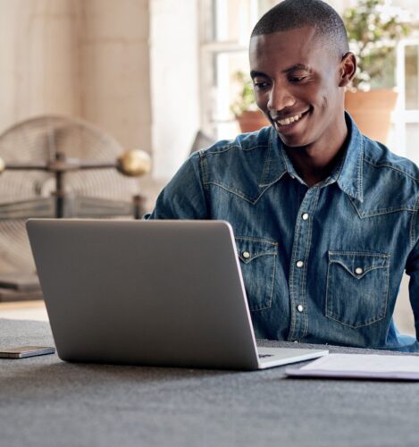 A person wearing a denim shirt sits at a table, smiling while using a laptop. Papers and a smartphone are on the table. A fan and potted plants are in the background.