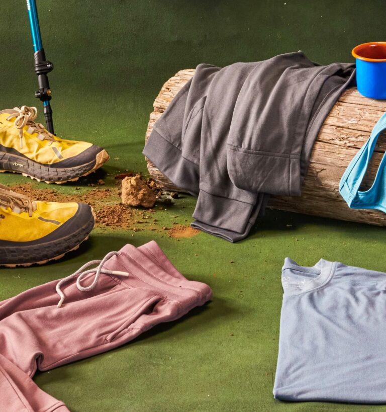Outdoor clothing and gear, including yellow shoes, a walking stick, a blue mug, and various clothing pieces, are arranged on artificial grass with a log in the background.