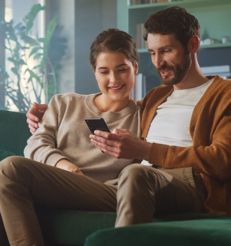 A man and woman sit on a green couch, smiling while looking at a smartphone together in a living room setting.